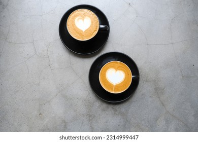 Two black mugs of cappuccino with heart shaped latte art on concrete background.