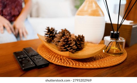 Two black remote controls sit beside pine cones and a reed diffuser on a table.