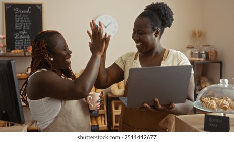 Two black women bakers high-fiving in a bakery shop, one holding a laptop and the other a coffee cup, displaying joyful and collaborative team spirit in an indoor setting - Powered by Shutterstock