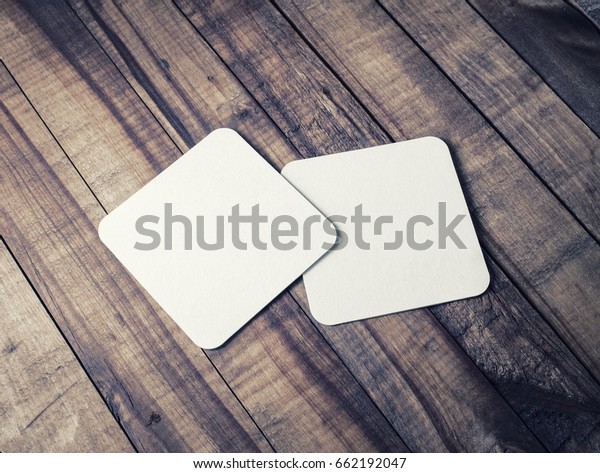 Two\
blank square beer coasters on wood table\
background.