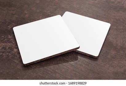 Two blank white quadratic drink coasters lying on the wooden table. Mock up. Clean square can mat isolated on a wooden table.