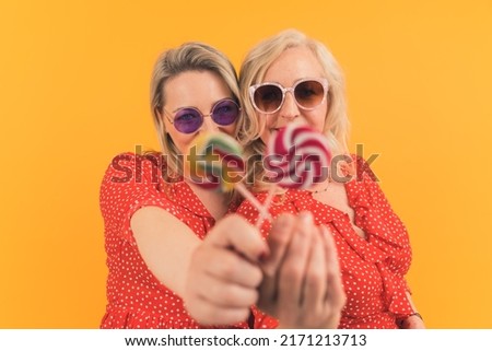 Two blonde cute middle-aged women wearing red outfits and funny sunglasses showing lollipops to camera. Focus on the background. High quality photo