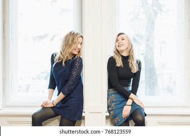 Two Blonde Female Friends Chatting And Laughing While Sitting By The Wall Between Two Windows. Breaking Down The Barrier Between People Concept