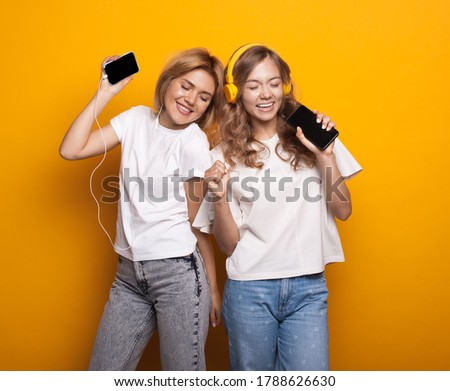 Two blonde women listening to music with headphones and mobile on yellow studio wall