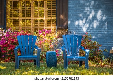 Two Blue Adirondack Chairs And Matching Drum Table Sitting On Lawn In Front Of Shuttered Window In Blue Brick House Landscaped With Azaleas In Dappled Light