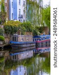 Two blue and brown wooden narrowboats or houseboats docked and moored in the residential district of Maida Vale, on the promenade below the typical Victorian English houses.