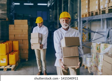 Two Blue Collar Workers In White Uniforms And With Yellow Helmets On Heads Relocating Heavy Boxes In Warehouse. Selective Focus On Senior Adult Employee.