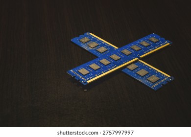 Two blue computer RAM memory sticks crossed to form an X shape. The modules are resting on a dark wood grain background, showcasing the black memory chips and gold contact pins. This image represents computer hardware components, DIY PC building, and is highly relevant for editorial topics such as the 2026 global RAM shortage, semiconductor supply chain issues, and hardware price inflation.