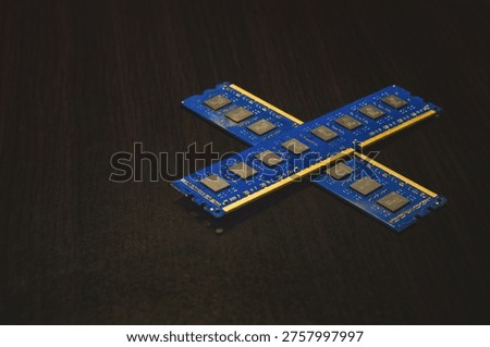 Two blue computer RAM memory sticks crossed to form an X shape. The modules are resting on a dark wood grain background, showcasing the black memory chips and gold contact pins. This image represents computer hardware components, DIY PC building, and is highly relevant for editorial topics such as the 2026 global RAM shortage, semiconductor supply chain issues, and hardware price inflation.