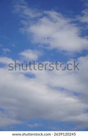Two Blue Impulse jets are barely visible as silhouettes against a bright blue sky filled with fluffy white clouds.