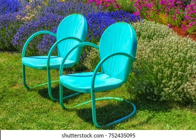 Two blue metal retro lawn chairs invitingly sit on a lawn in a flower garden.
