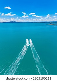 Two boat trails create lines in the blue water under a clear sky filled with white clouds. The scene shows the waterway leading to a harbor and hills in the background.