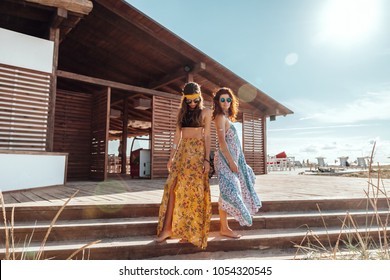 Two boho friends (girls) wearing floral maxi dress and skirt relaxing on the beach. Bohemian clothing style.
