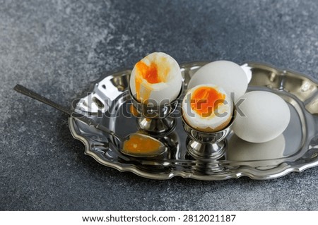 Two boiled eggs are in silver egg cups one with a soft yolk. A spoon rests beside the cups on a silver tray. The background is gray and simple.