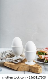 Two boiled eggs, whole in shell, in white egg stand, on wooden Board, textile napkin, toast with bacon, light grey background, space for text