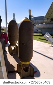 Two bombs stand on display at a military aviation museum. The bombs feature a yellow tip and are placed next to various aircraft. Visitors can be seen in the background.