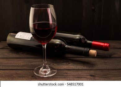 Two Bottles Of Red Wine With A Glass On An Old Wooden Table. Focus On The Stem Glass