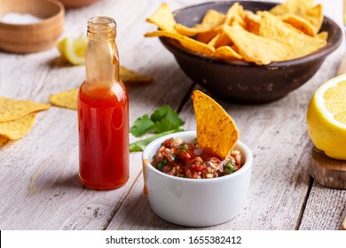 Two Bowls Full Of Salsa Dip, Hot Red Chili Sauce Bottle And Tortilla Chips On Rustic Wooden Table, Sause Being Added To One Of The Bowls