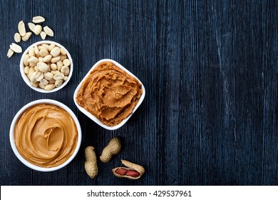 Two bowls of peanut butter and peanuts on dark wooden background from top view