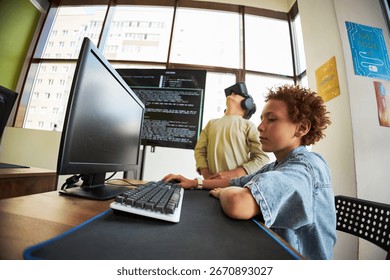 Two boys in classroom using technology, Caucasian child sitting at desk typing on computer keyboard while another child wearing virtual reality headset standing in background