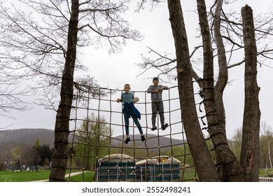 Two boys climbing on rope net obstacle course between trees on cloudy day in the park. Concept of outdoor adventure, teamwork, and active childhood fun in nature. High quality photo - Powered by Shutterstock