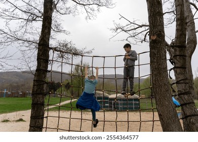 Two boys climbing on rope net playground structure between trees on an overcast day in park. Concept of teamwork, adventure, outdoor activities, and childhood fun in nature. High quality photo - Powered by Shutterstock