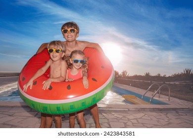 Two boys and a girl in swimwear. sunglasses pose with a large watermelon inflatable by a pool, against a backdrop of the setting sun. - Powered by Shutterstock