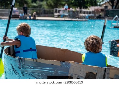 Two Boys Paddling A Cardboard Boat In A Race In A Pool