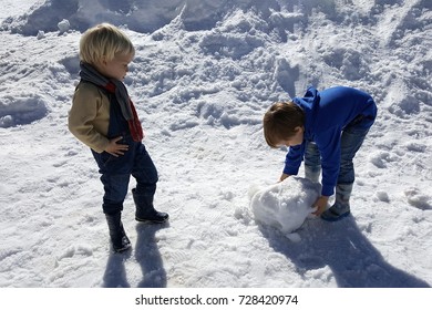 Two Boys Play In The Winter On Snow. 3 Years Child Hands In Pants Is Looking On 6 Years Kid Bends Down Is Working, Trying To Lift.