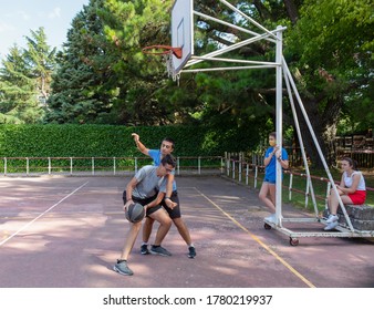 Two Boys Playing Basketball On An Outdoor Basketball Court And Two Girls Watching Them Wearing Masks In Time Of Covid19