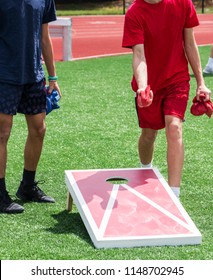 Two Boys Are Playing A Game Of Corn Hole During A High School Gym Class On The Turf.