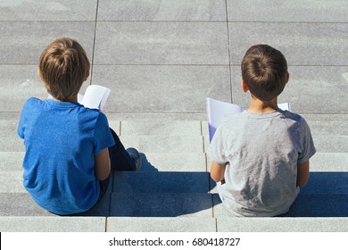 Two boys reading books sitting on the stairs outdoors, back view