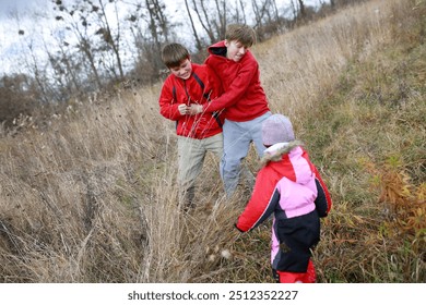 Two boys in red jackets engage in playful wrestling in a grassy field while a young girl in winter attire giggles and watches them. The atmosphere is cheerful and energetic. - Powered by Shutterstock