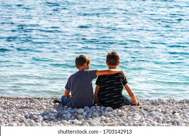 Two boys sitting on the beach, talking and throwing rocks into the sea