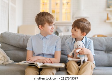 Two boys sitting on couch are reading book. Older brother reads interesting stories to his younger brother, concept friendship, intelligence, interaction, younger brother holds toy rabbit in his hand - Shutterstock ID 2117845397