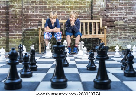 Two boys, sitting on a wooden bench, concentratedly thinking about their next move during an outdoors chess game with life sized pieces.