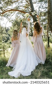 Two bridesmaids in powdery dresses holding with bouquets in hands stand with their backs near the bride in a white dress with a wedding bouquet in her hand on a green lawn