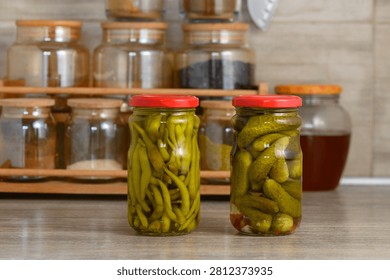 Two bright jars hold delicious pickled green beans and cucumbers, showcasing vibrant colors and textures. The kitchen background features a wooden shelf filled with various jars, adding a cozy, homey atmosphere to the scene.