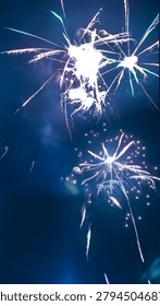 Two bright white fireworks exploding in a dark blue night sky, one dense like a sparkler, the other daisy-shaped with light trails, surrounded by smoke.
