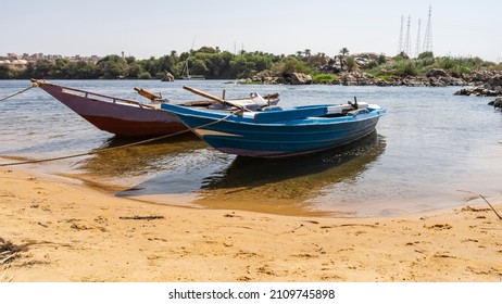 Two bright wooden boats are tied up near the bank of the Nile. There is yellow sand on the beach. Shadows and reflections on clear water. Green vegetation, boulders against the blue sky. Egypt. Aswan