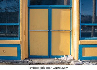 Two Bright Yellow Doors With Chrome Handle To A Store With Blue Trim. Over The Door Is A Transom Glass Window With Multiple Panes. There's A Sidewalk In Front Of The Tall Entrance To The Vintage Shop 