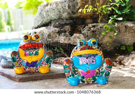 Two brightly colored and decorated ceramic frogs sit by landscaping rocks by swimming pool