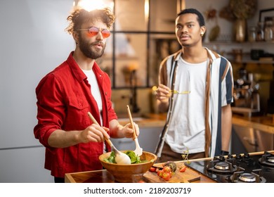 Two Brightly Dressed Guys Having Fun While Making Salad Together On Kitchen. Concept Of Gay Couples And Everyday Life At Home. Caucasian And Hispanic Man Cooking Healthy Food