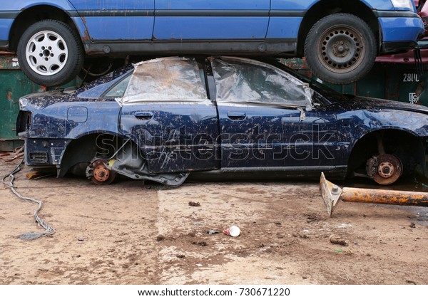 Two broken blue cars, one on top of\
the other, in a scrap yard. One car without wheels and broken\
windows. Plastic covering the windows. Mud over the\
cars.