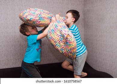 Two Brothers Of A European Boy Fight With Pillows On The Bed. A Merry Duel Of Children. Fight At Home Between Friends, War On Purpose.