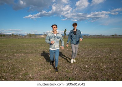 Two Brothers Run In A Field Of Grass In The Open Air, Young Men Dressed Causally, Wearing Denim Jackets. Brotherhood Concept.