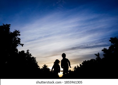 Two Brothers Run Together Through A Field Between Trees, At Sunset In Silhouette.