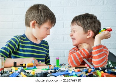 Two Brothers Sit At The Table And Assemble The Designer, Build From Toy Cubes. One Child Laughs, The Other Looks Serious. Fun Collaborative Activity. The Boys Are Friends, Busy With A Common Cause