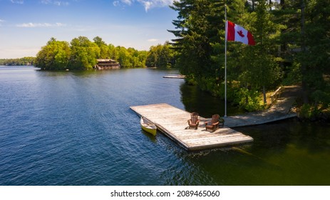Two brown Adirondack chairs on a wooden pier with a yellow canoe. Across the calm water is a brown cottage nestled among green trees. Canada flag is waving on a pole.