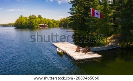 Two brown Adirondack chairs on a wooden pier with a yellow canoe. Across the calm water is a brown cottage nestled among green trees. Canada flag is waving on a pole.